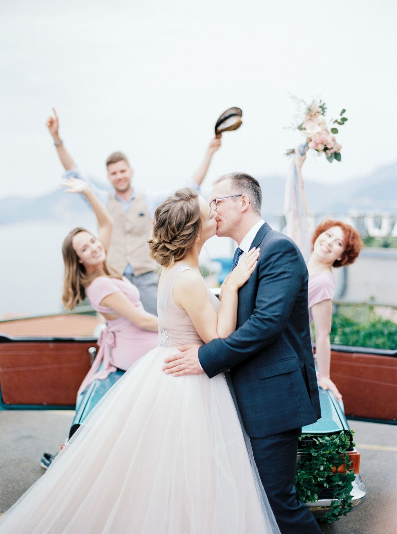 Bride and groom kissing on a seaside terrace while friends cheer and wave a hat