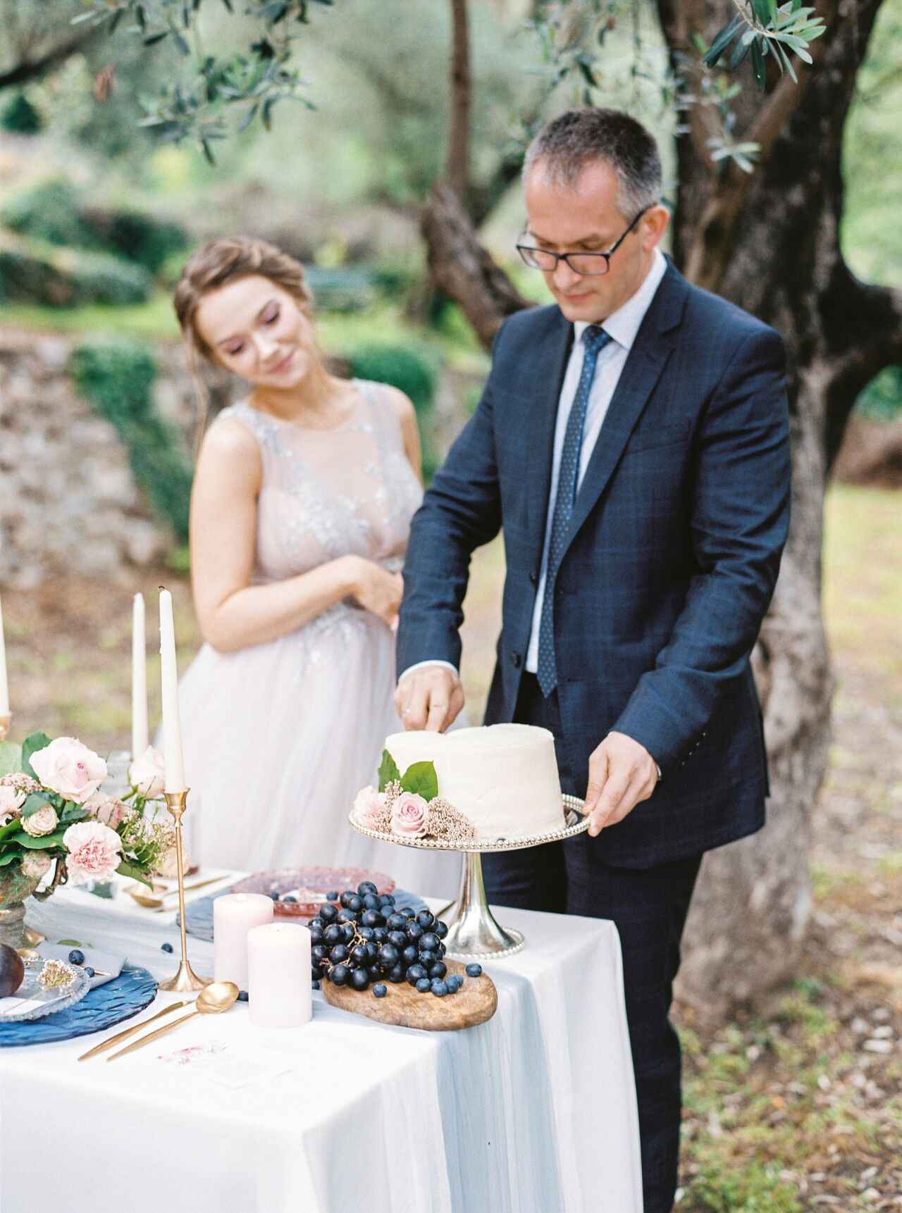 Bride and groom cutting a white rose-topped cake at an outdoor table set with grapes and candles