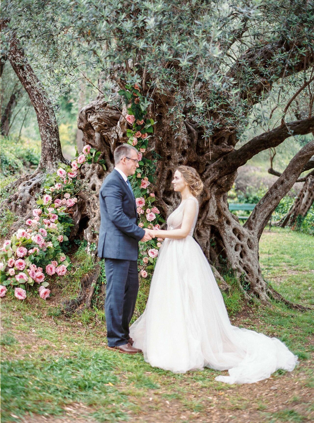 Bride and groom holding hands for their vows under a rose-covered olive tree