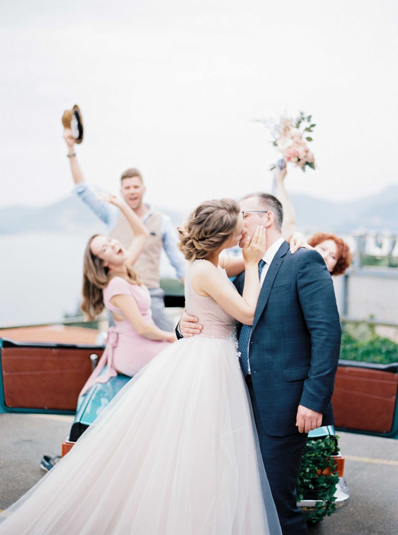Bride and groom kissing as friends cheer behind them on a terrace above the sea