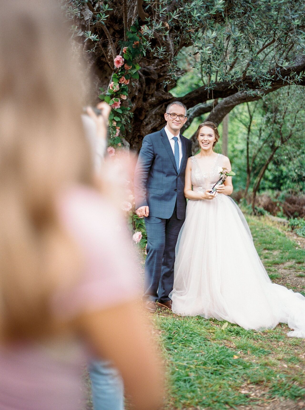 Bride and groom smiling under an olive tree, seen past a guest in the foreground