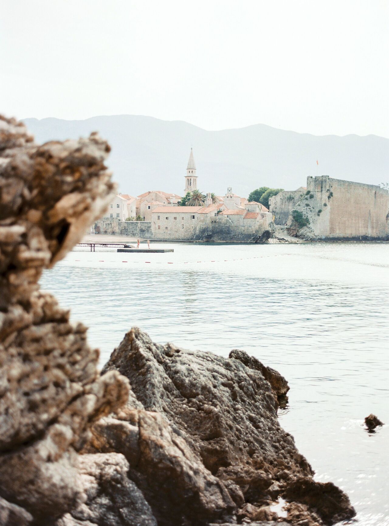 Budva's old town walls seen past dark coastal rocks across still water