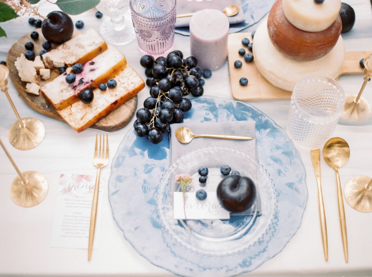 Overhead view of a reception place setting with a blue glass plate, gold cutlery, figs and grapes