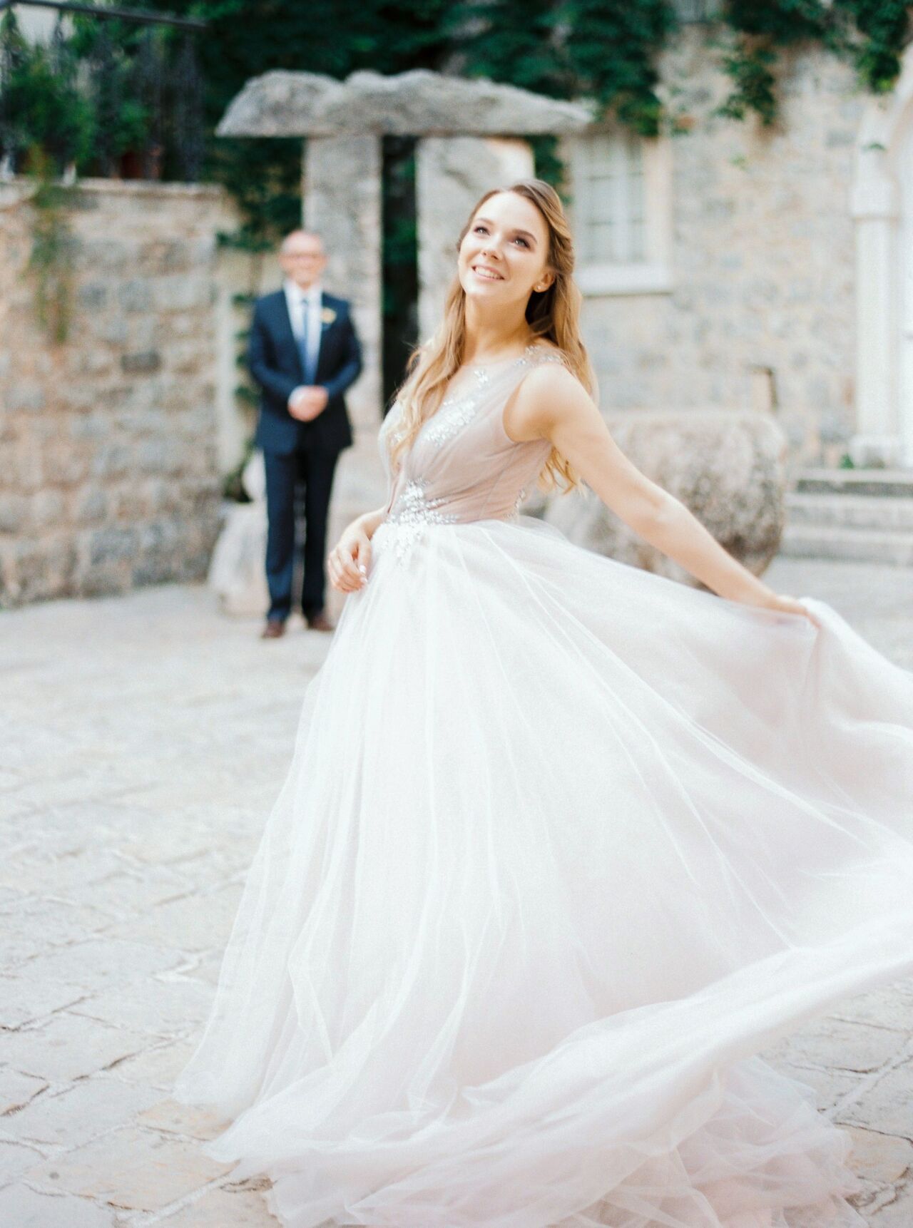 Bride twirling her full tulle skirt in a sunlit stone courtyard while the groom watches